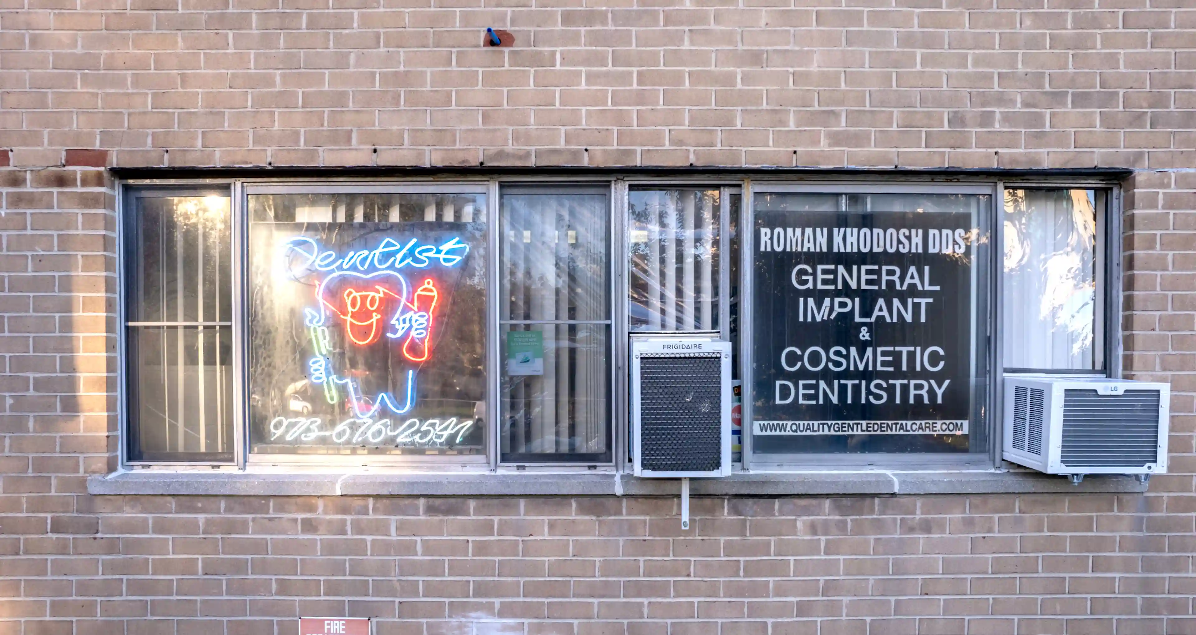 Exterior brick wall with three windows, one featuring a neon dentist sign, and another with a sign for Roman Khodosh DDS general implant and cosmetic dentistry.